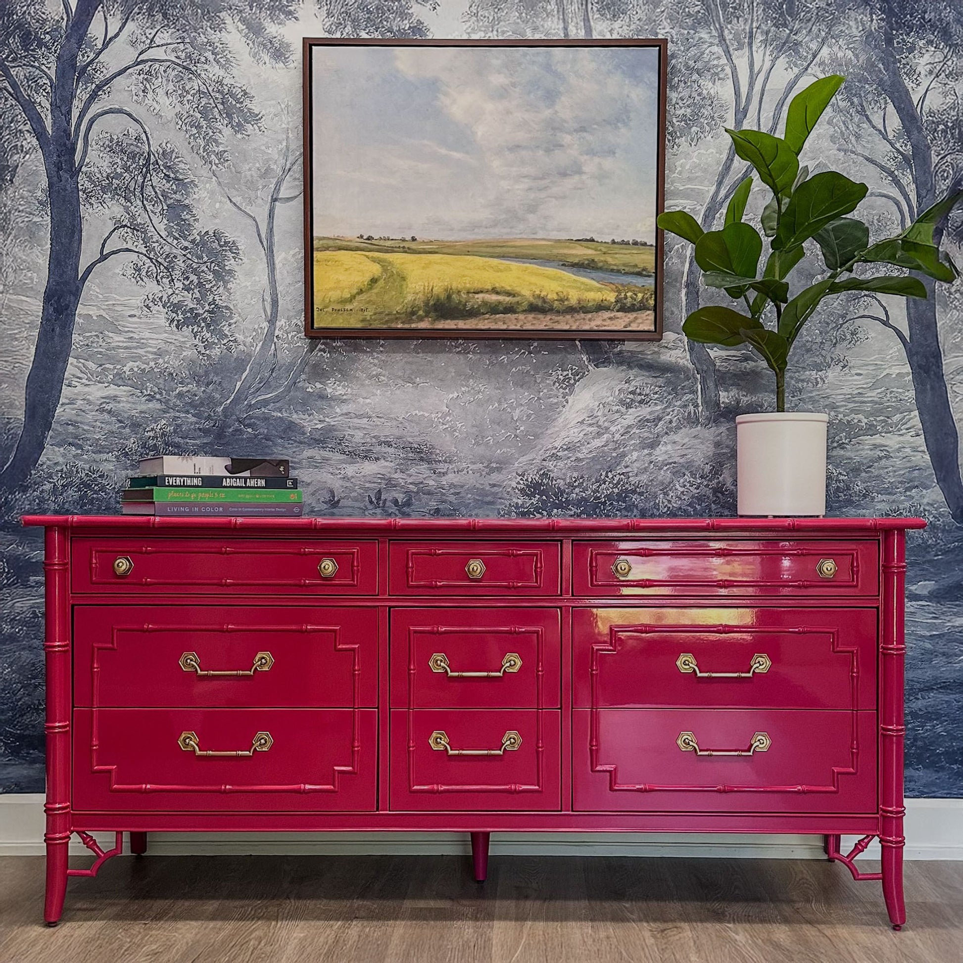 Red dresser with gold handles against a wall with a landscape painting and plant.